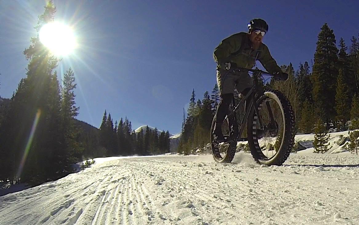 A person riding a fat bike on a snowy trail surrounded by pine trees, with the sun shining brightly in the sky. Snow is being kicked up from the bike's tires, creating a dynamic outdoor scene. Cottonwood Pass Road mountain bike trail.