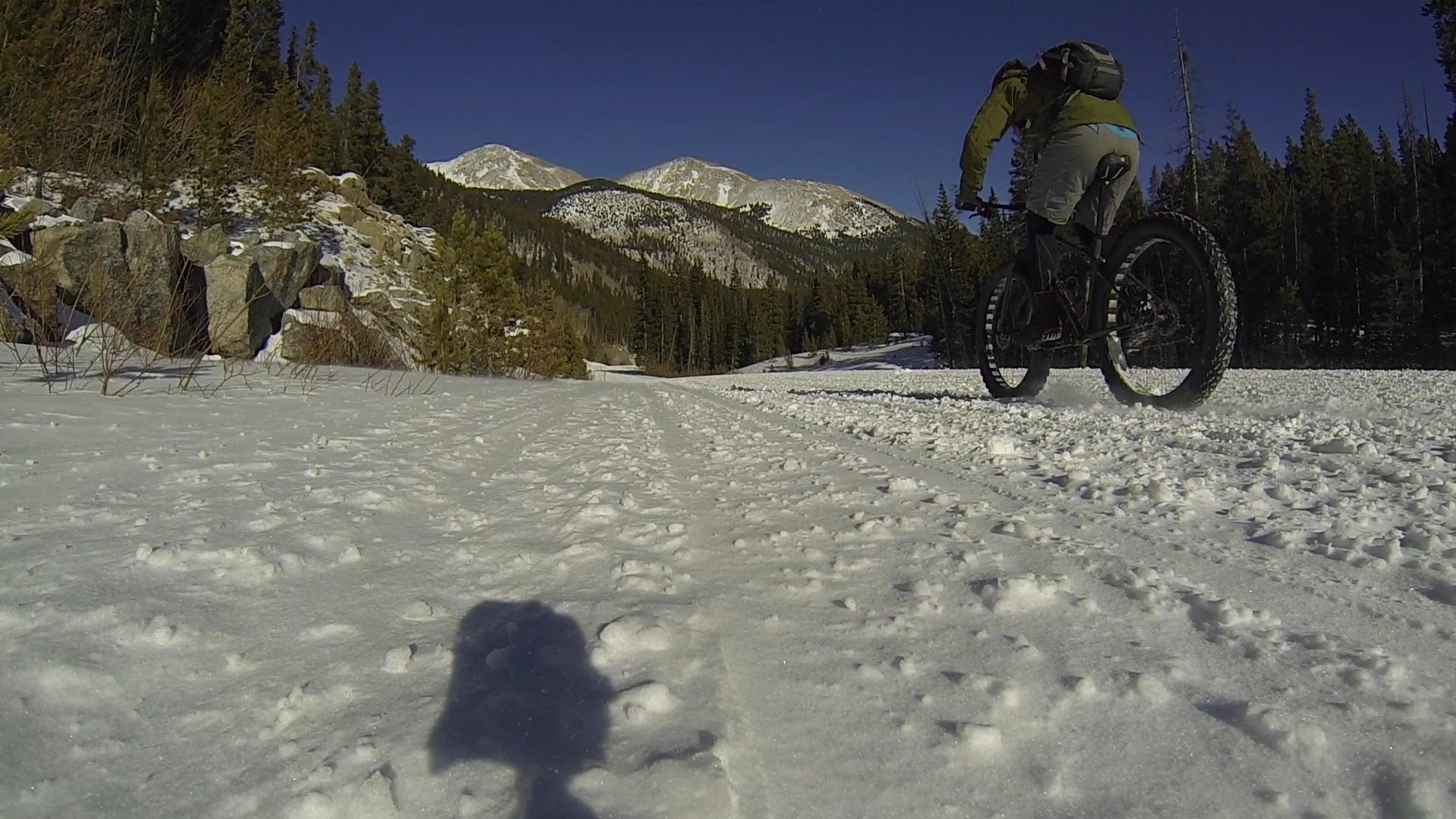 A person riding a fat bike on a snowy trail, with a mountainous landscape in the background and a clear blue sky. The foreground shows uneven, snow-covered terrain, while the surrounding area is filled with evergreen trees. Cottonwood Pass Road mountain bike trail.