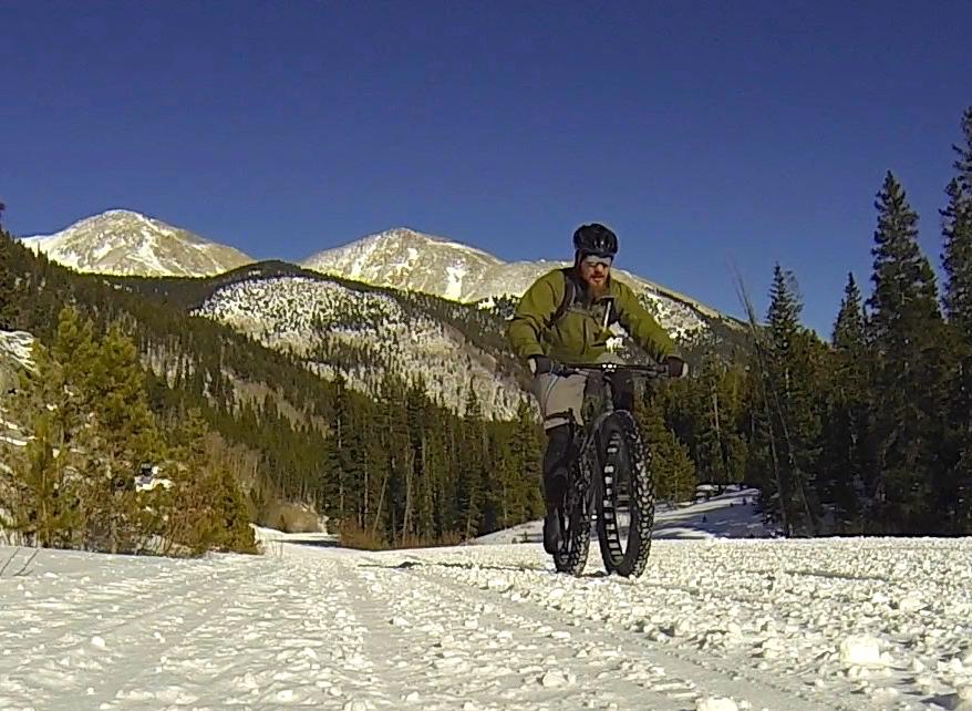 A person riding a fat bike on a snowy trail surrounded by trees and mountains under a clear blue sky. Cottonwood Pass Road mountain bike trail.