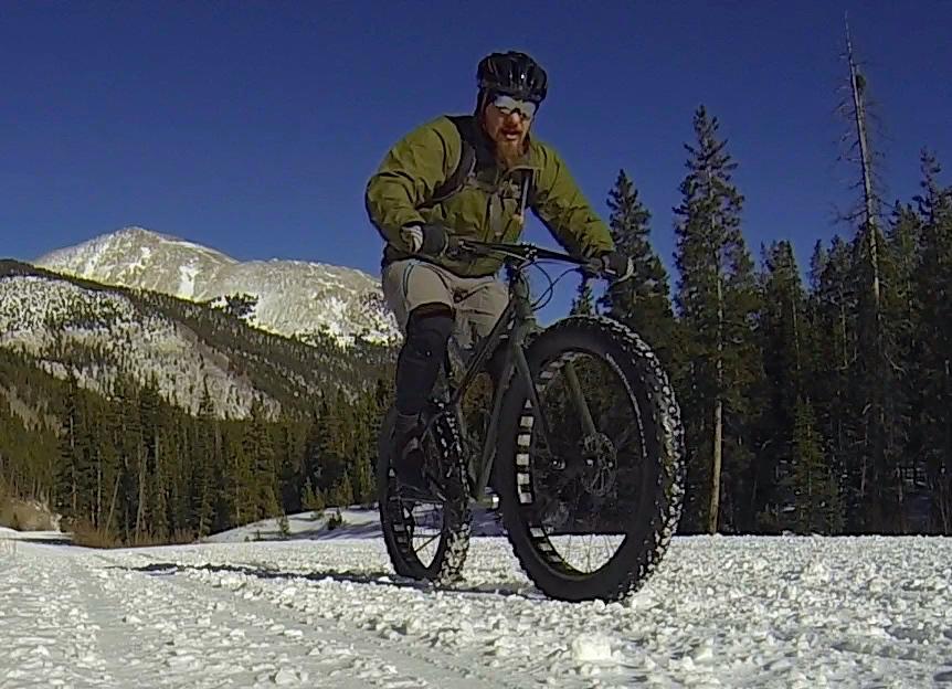A person riding a fat bike on a snowy trail, surrounded by tall coniferous trees and mountains in the background under a clear blue sky. The cyclist is wearing a green jacket and has a beard, focused on navigating the winter terrain. Cottonwood Pass Road mountain bike trail.