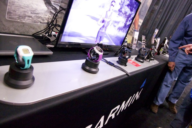 A display of various Garmin smartwatches on a promotional table, featuring multiple models in different colors mounted on stands. A television in the background shows a video of a person jogging outdoors. Some attendees are interacting near the display.