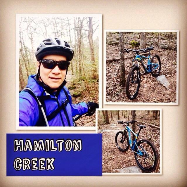 A biker in a blue jacket and black helmet poses for a selfie on a wooded trail, surrounded by trees. The image also features two additional photos: one of the biker’s blue mountain bike parked on a trail, and another close-up of the bike. The text "HAMILTON CREEK" is prominently displayed at the bottom of the image. Hamilton Creek mountain bike trail.