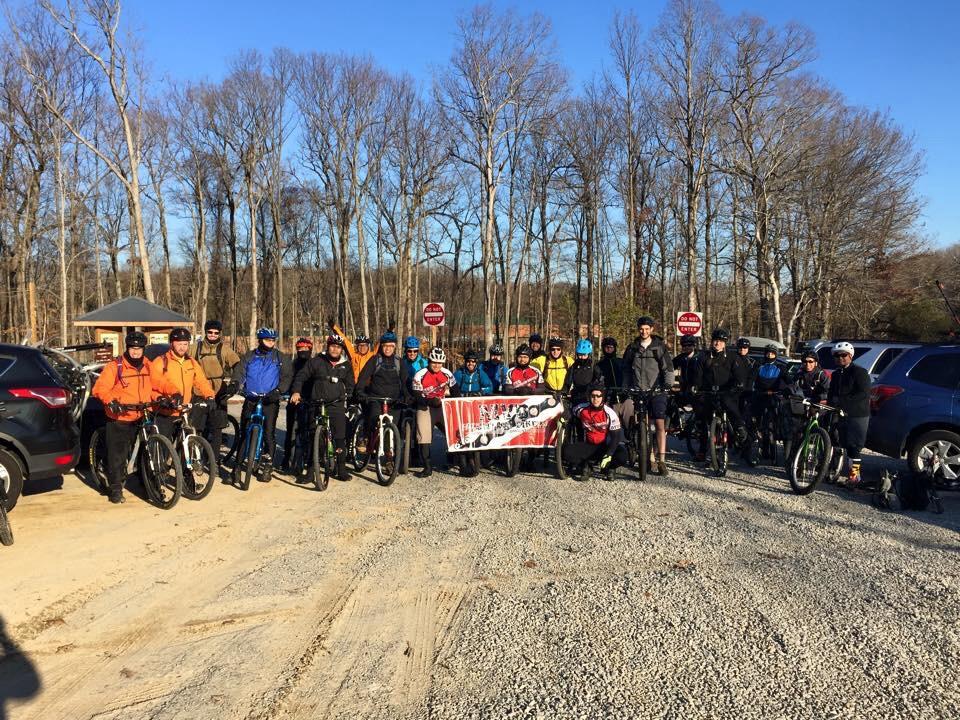 A group of diverse individuals stands in front of a banner, gathered with their mountain bikes in a gravel parking area surrounded by bare trees. The participants are dressed in various outdoor clothing, suggesting a chill day for biking. Some individuals are wearing helmets and appear excited to embark on a ride, with a few parked cars visible in the background. Meadowood mountain bike trail.