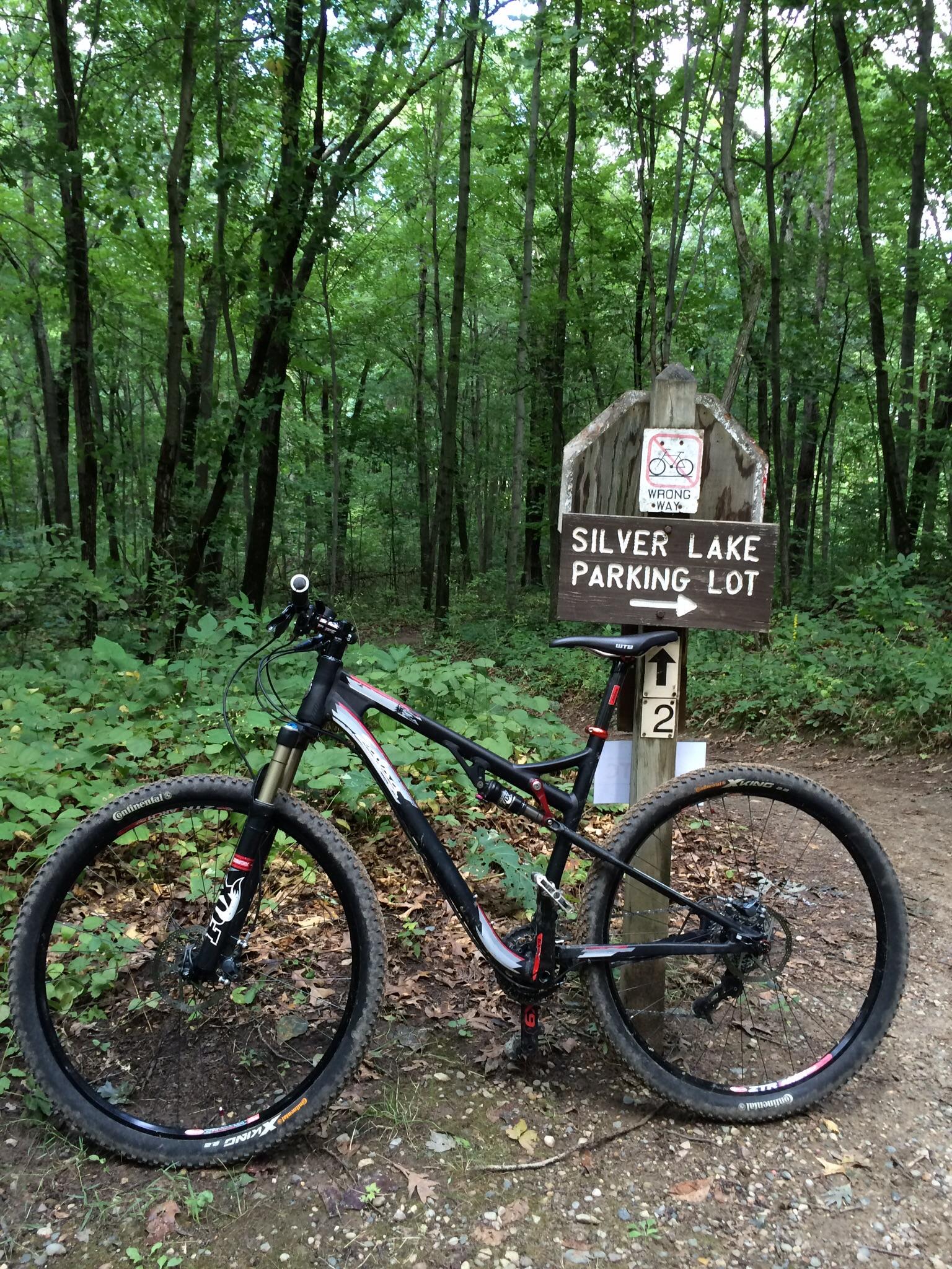 A mountain bike parked next to a wooden sign that reads "Silver Lake Parking Lot," surrounded by dense greenery and trees. The area is a natural trail setting, indicating a location popular for biking and outdoor activities. Potawatomi trail mountain bike trail.