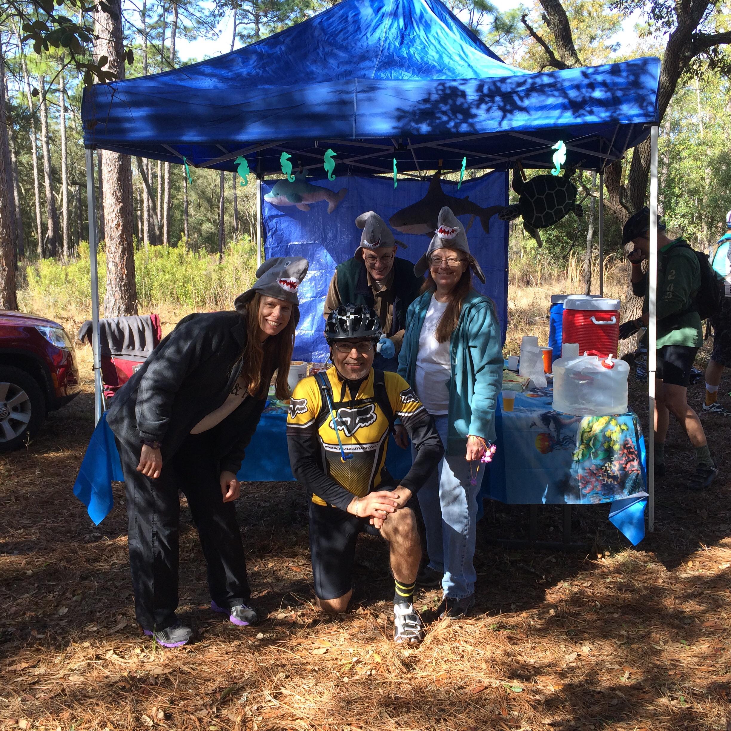 A group of four people wearing shark-themed hats poses together in front of a decorated canopy at an outdoor event. One person, dressed in a bike jersey and helmet, kneels in the foreground, while the others stand behind them, all smiling. The backdrop features a blue fabric with marine motifs, including artificial sharks and sea turtles. Pine trees and a car are visible in the background, indicating a wooded setting. Withlacoochee State Forest: Croom Section mountain bike trail.