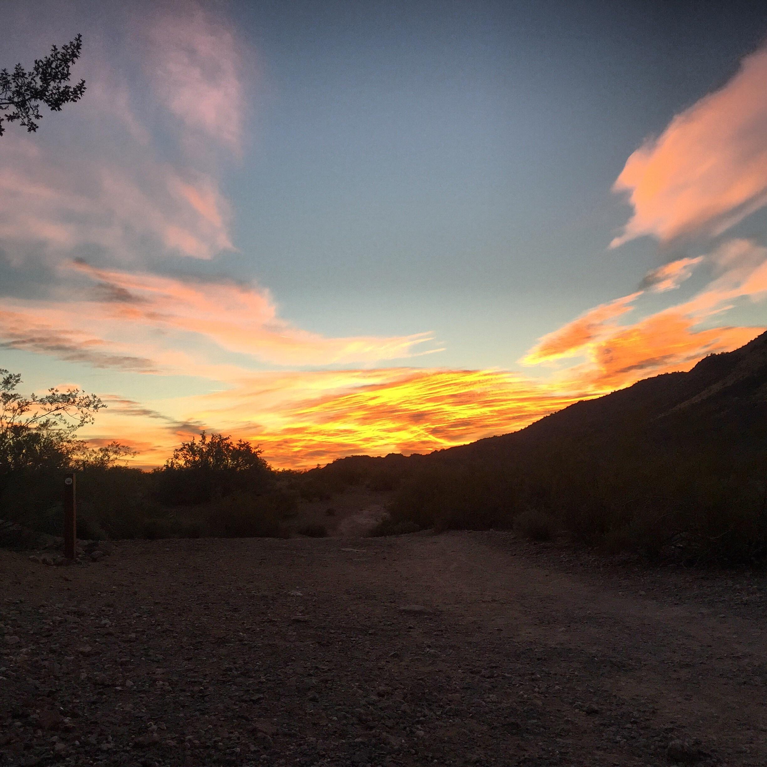 A scenic sunset over a desert landscape, featuring vibrant orange and pink clouds in the sky, with silhouettes of mountains and sparse vegetation in the foreground. The scene conveys a tranquil and picturesque evening atmosphere. Trail #100 mountain bike trail.