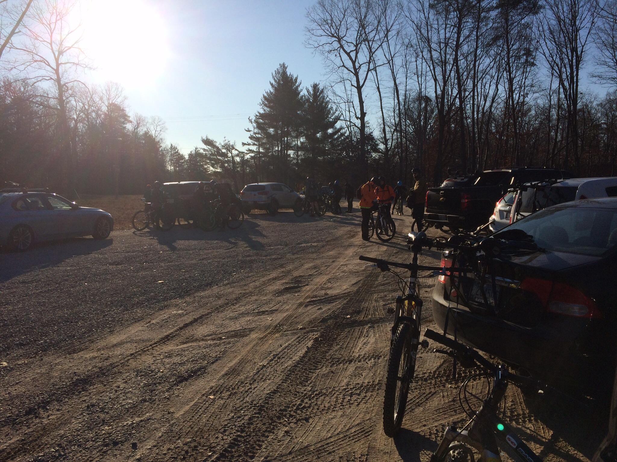 A gravel parking lot filled with cars and bicycles, set against a backdrop of trees during a sunny day. Several people are gathered, preparing for a biking outing, with some wearing bright clothing and others on bicycles. The sun shines brightly in the sky, illuminating the scene. Meadowood mountain bike trail.