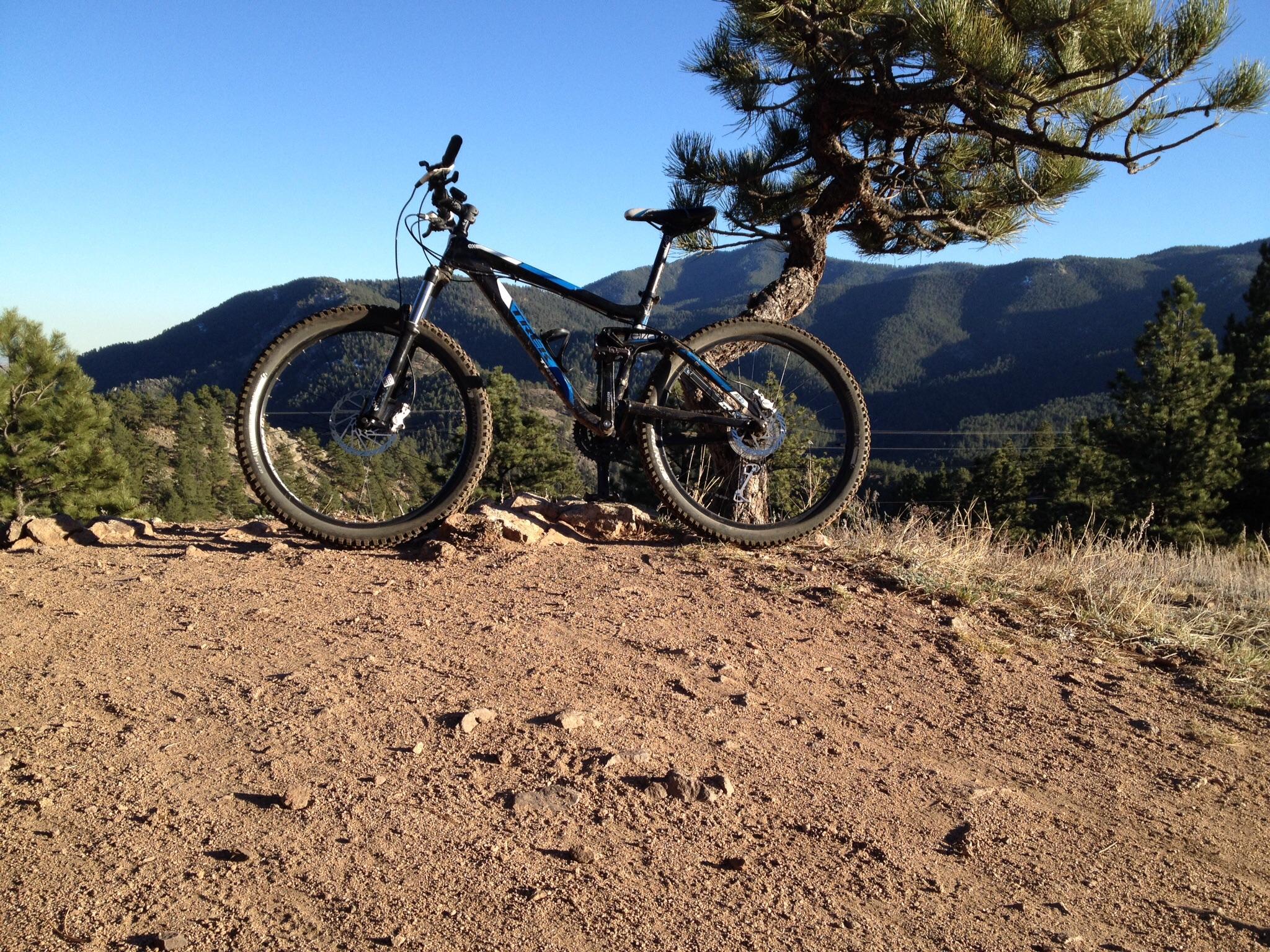 Trek Fuel EX 5: A mountain bike parked on a dirt path, with green trees and rocky hills in the background under a clear blue sky.