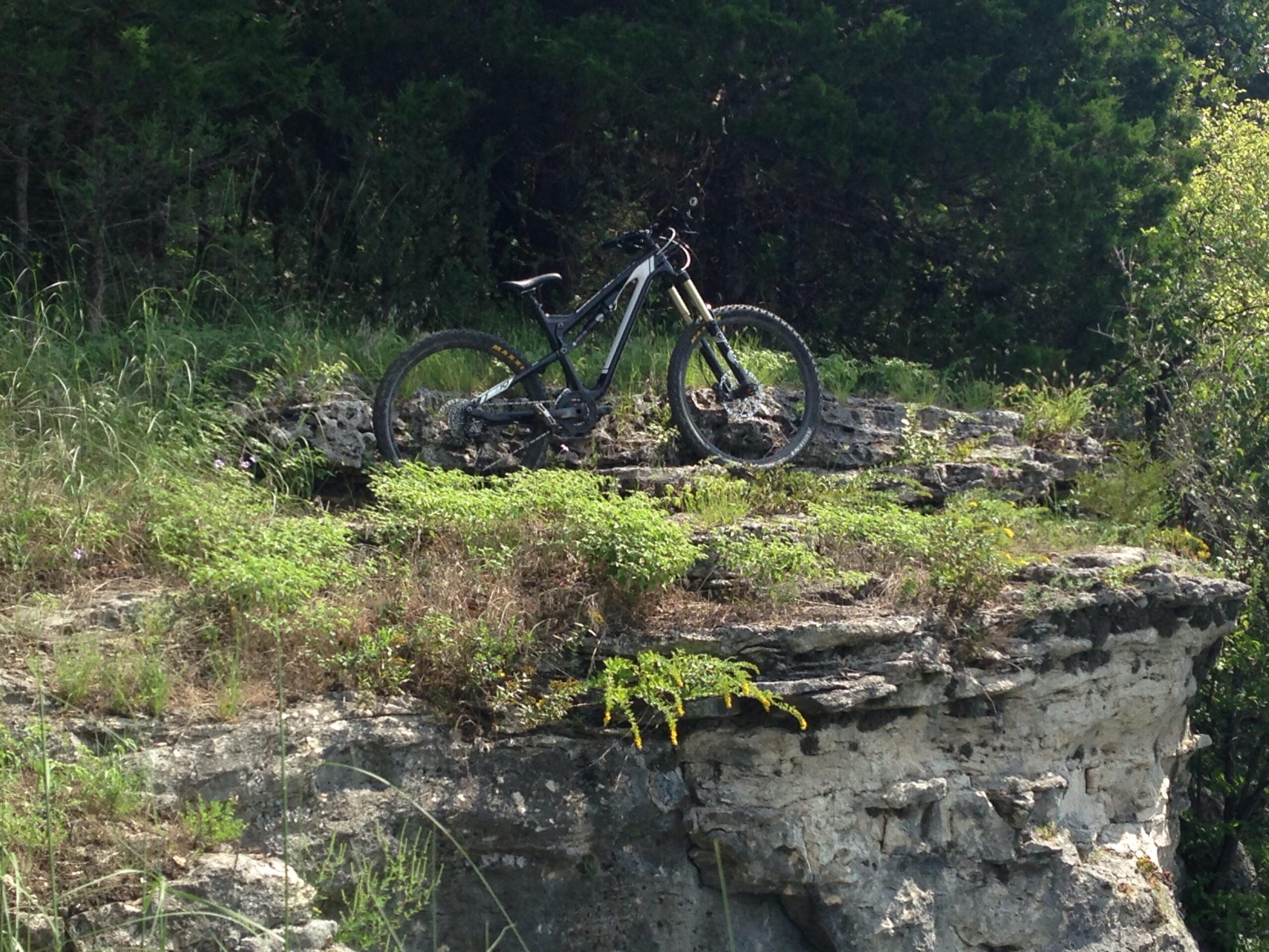 Scott Scott genius lt 710: A mountain bike resting on a rocky ledge surrounded by lush green vegetation under a clear sky.