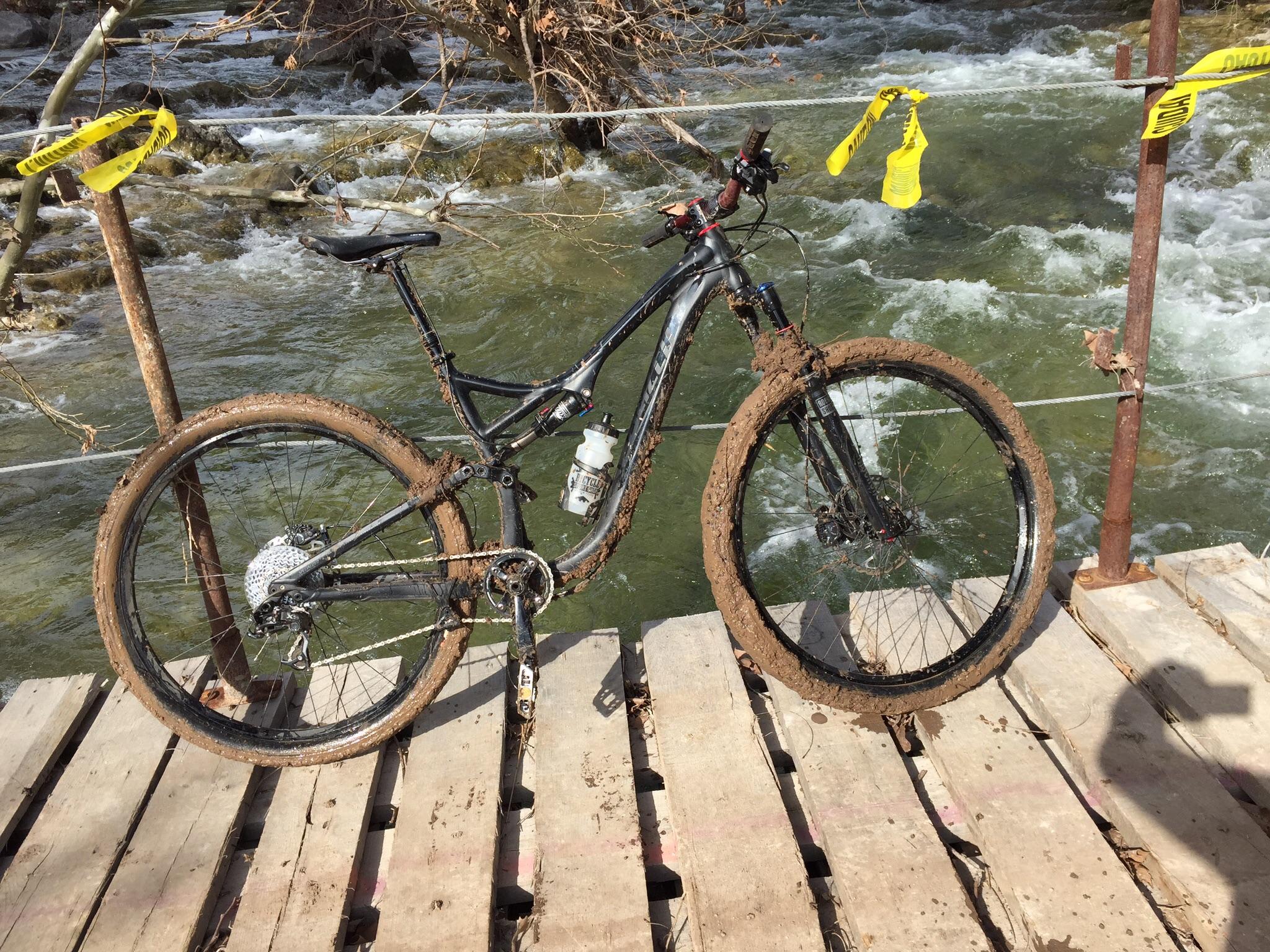 A mountain bike covered in mud is resting on a wooden bridge over a flowing stream. Yellow caution tape is visible in the background, indicating a potential hazard nearby. The bike's tires are particularly muddy, suggesting it has recently been used in rugged outdoor terrain. Barton Creek Greenbelt mountain bike trail.