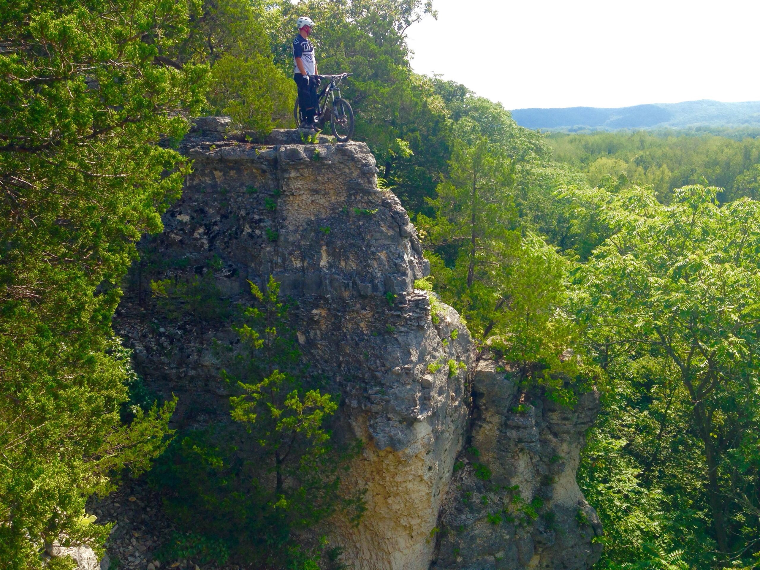 Scott Scott genius lt 710: A person standing on a rocky outcrop overlooking a lush green landscape, holding a mountain bike. The scene features trees and hills in the distance, with bright sunlight illuminating the area.