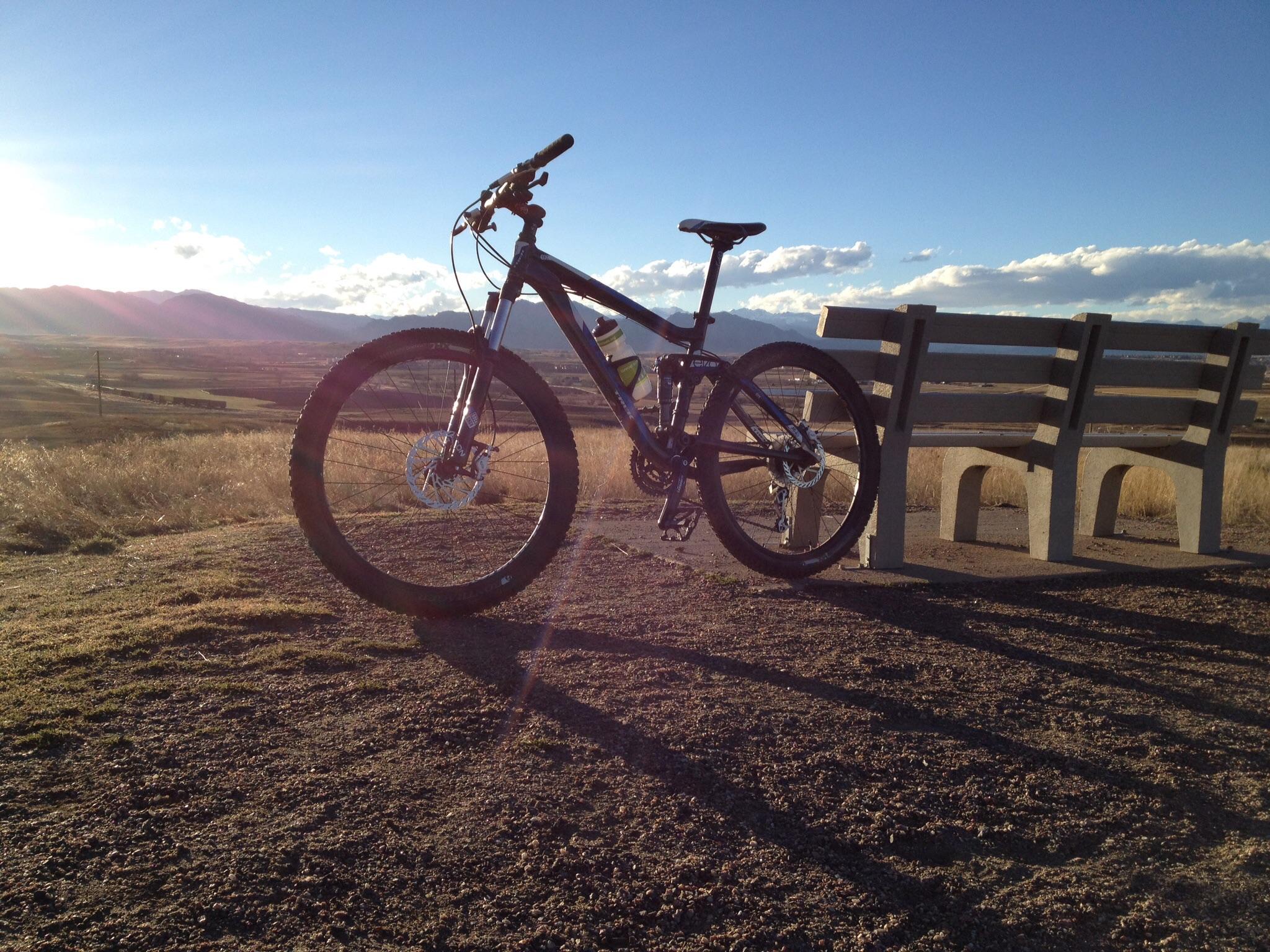 Trek Fuel EX 5: A mountain bike parked beside a concrete bench on a hilltop, with expansive views of rolling hills and mountains in the background during sunset. The scene is illuminated by warm sunlight, casting long shadows on the ground.