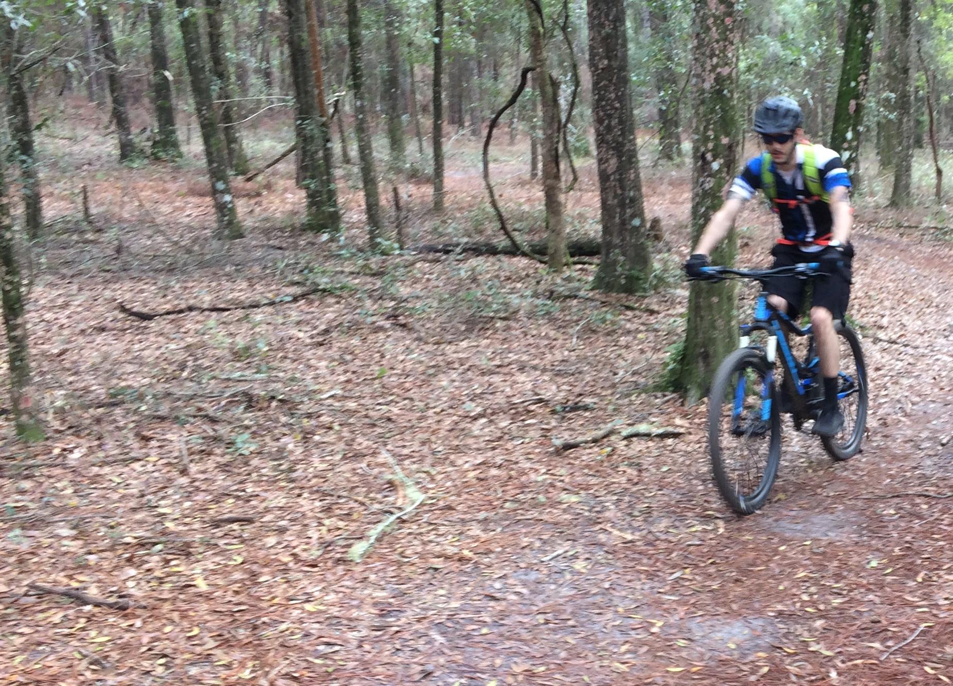 A person riding a mountain bike along a dirt trail in a wooded area, surrounded by trees and fallen leaves. The cyclist is wearing a helmet, sunglasses, and athletic clothing, contributing to an outdoor adventure atmosphere. Bunny mountain bike trail.