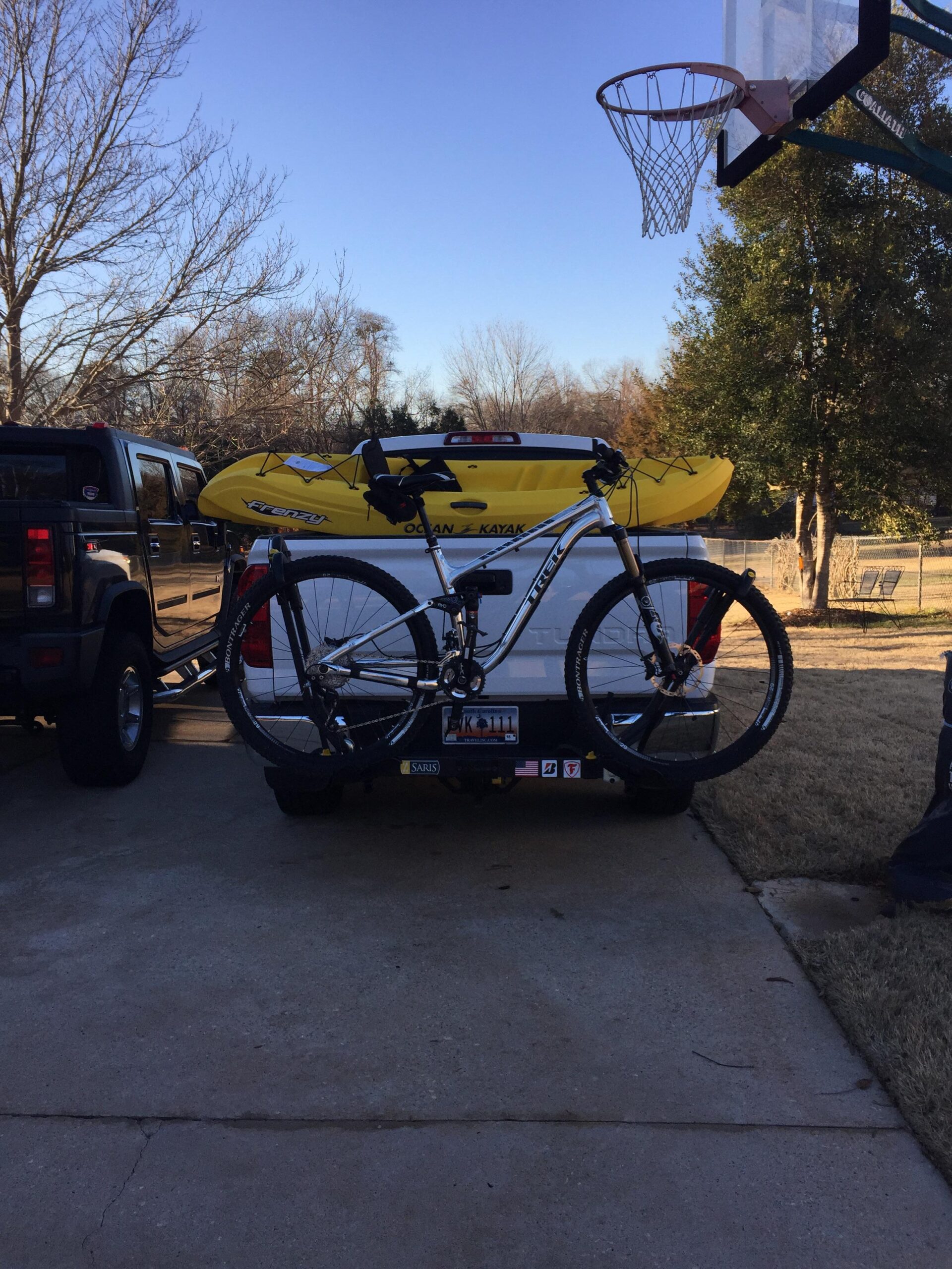Trek Fuel EX 9: A pickup truck parked in a driveway, with two yellow kayaks secured on the truck bed and a mountain bike attached to the back. In the background, there's a basketball hoop and trees, under a clear blue sky.