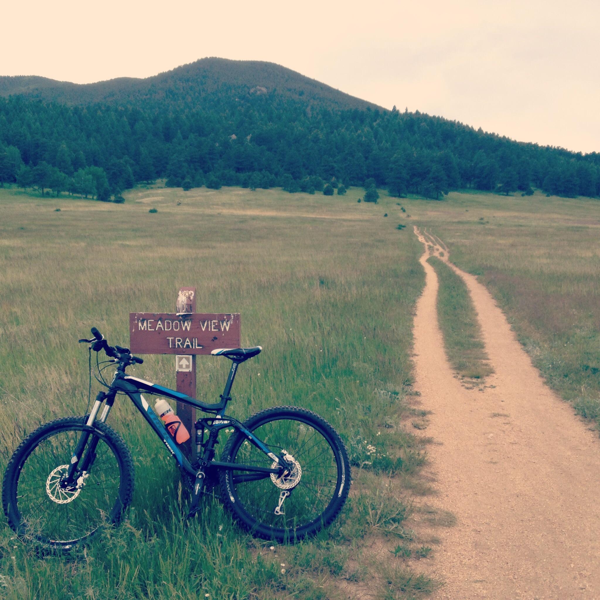 Trek Fuel EX 5: A mountain bike resting against a trail sign labeled "Meadow View Trail," with a dirt path leading away through a grassy field and rolling hills in the background. The scene is set in a natural landscape with trees and mountains under a cloudy sky.