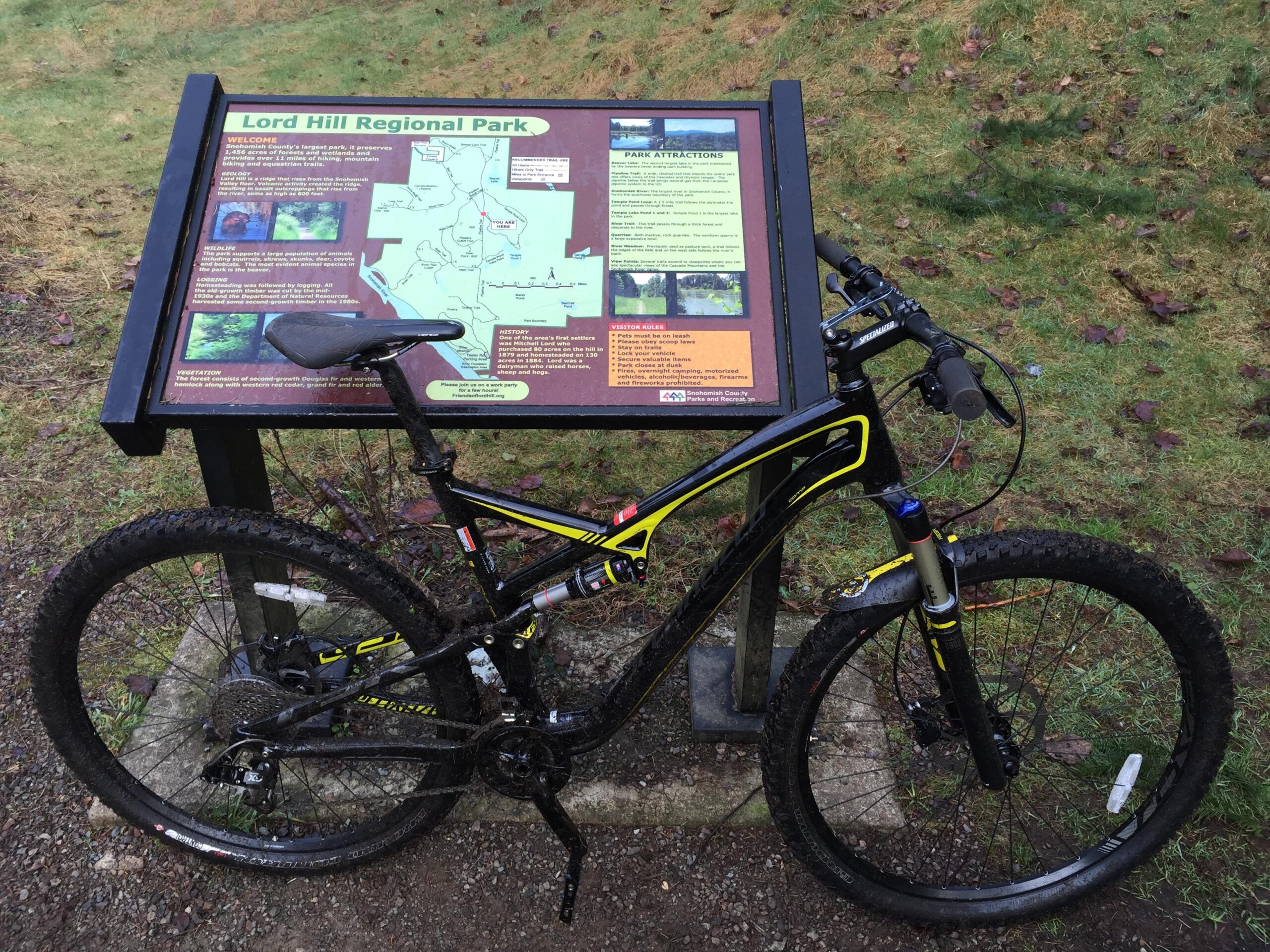 Specialized Camber Comp 29: A mountain bike parked next to a informational sign at Lord Hill Regional Park, showcasing a map and details about the park's attractions and guidelines. The ground is grassy with some fallen leaves, and the setting is slightly damp, indicating recent weather.