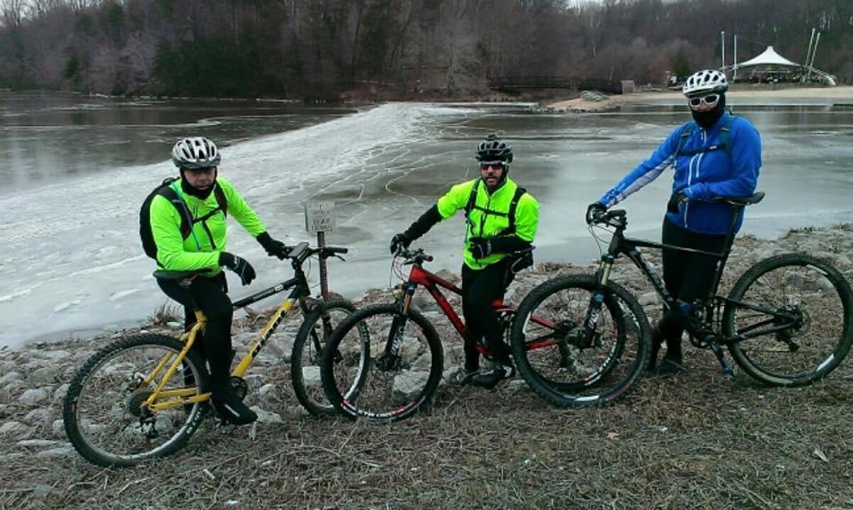 Three mountain bikers dressed in bright yellow and blue jackets pose alongside their bicycles near an icy lake. The background features a wooded area and a frozen shoreline, suggesting cool weather conditions. The bikers stand on a rocky bank, displaying their bikes while wearing helmets and warm clothing. Wakefield mountain bike trail.