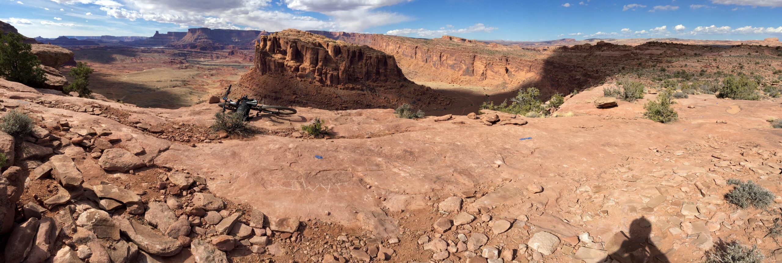 A panoramic view of a rugged landscape featuring red rock formations and canyons under a partly cloudy sky. A mountain bike lies on its side on a rocky surface, with scattered shrubs and vegetation visible. The vast expanse of the canyon extends into the distance, showcasing the natural beauty of the area. Captain Ahab mountain bike trail.