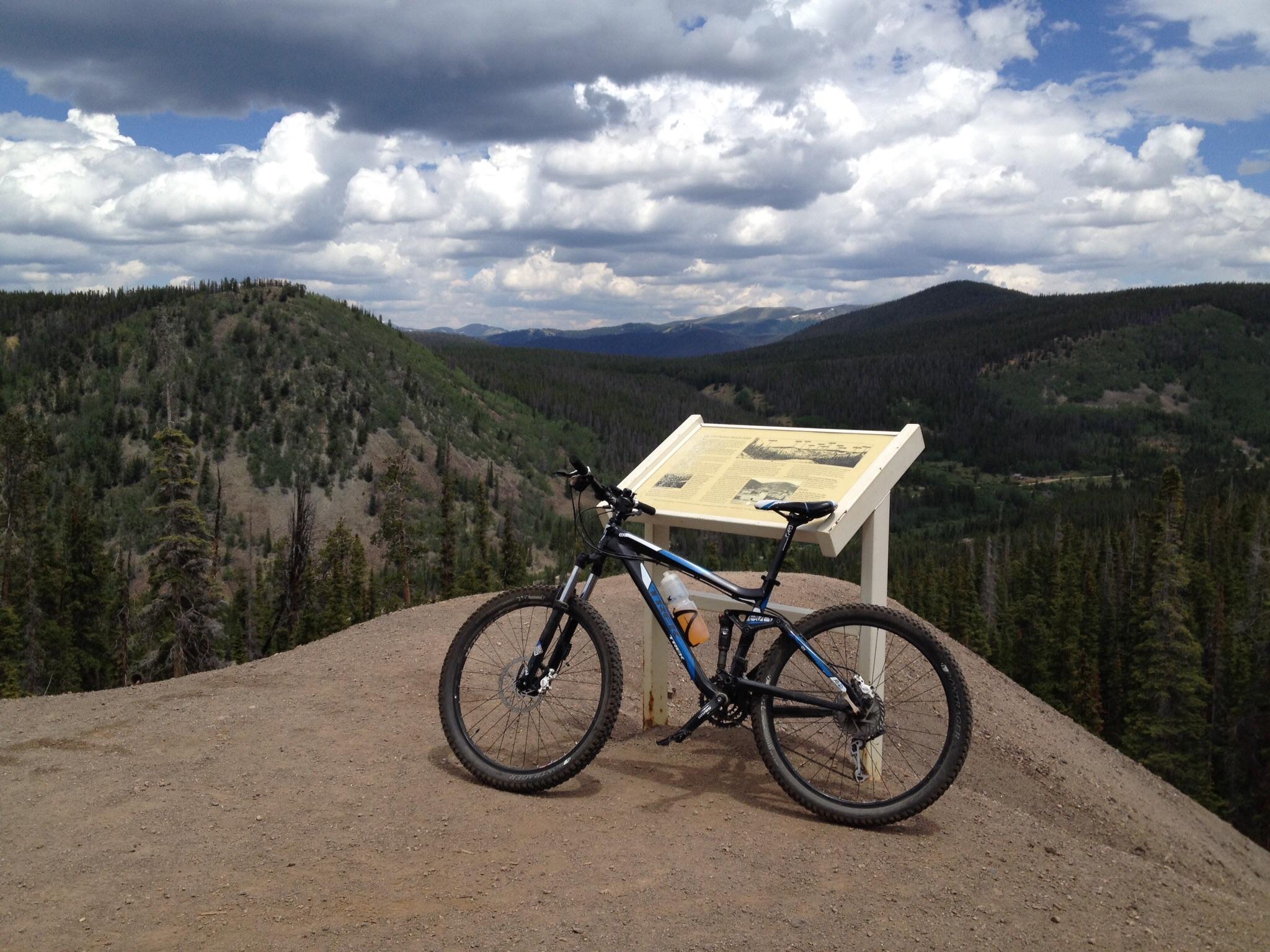 Trek Fuel EX 5: A mountain bike rests on a dirt path overlooking a lush, green landscape with rolling hills and a cloudy sky in the background. In the foreground, a sign with informational text is positioned nearby, providing context about the scenic area.