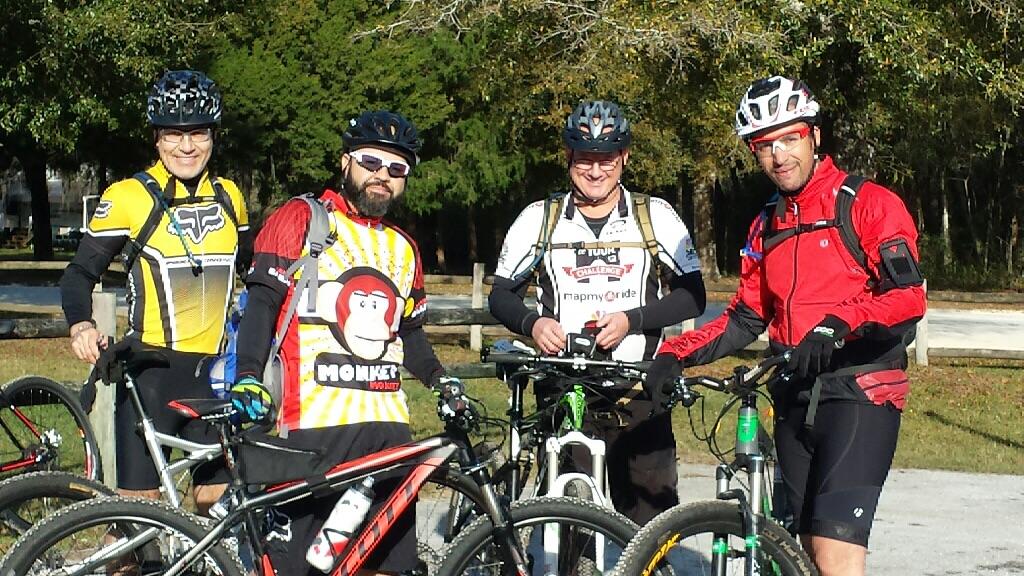 A group of four mountain bikers posing together outdoors, wearing colorful cycling jerseys and helmets, with their bikes beside them. The background features trees and a clear sky, indicating a sunny day. Withlacoochee State Forest: Croom Section mountain bike trail.