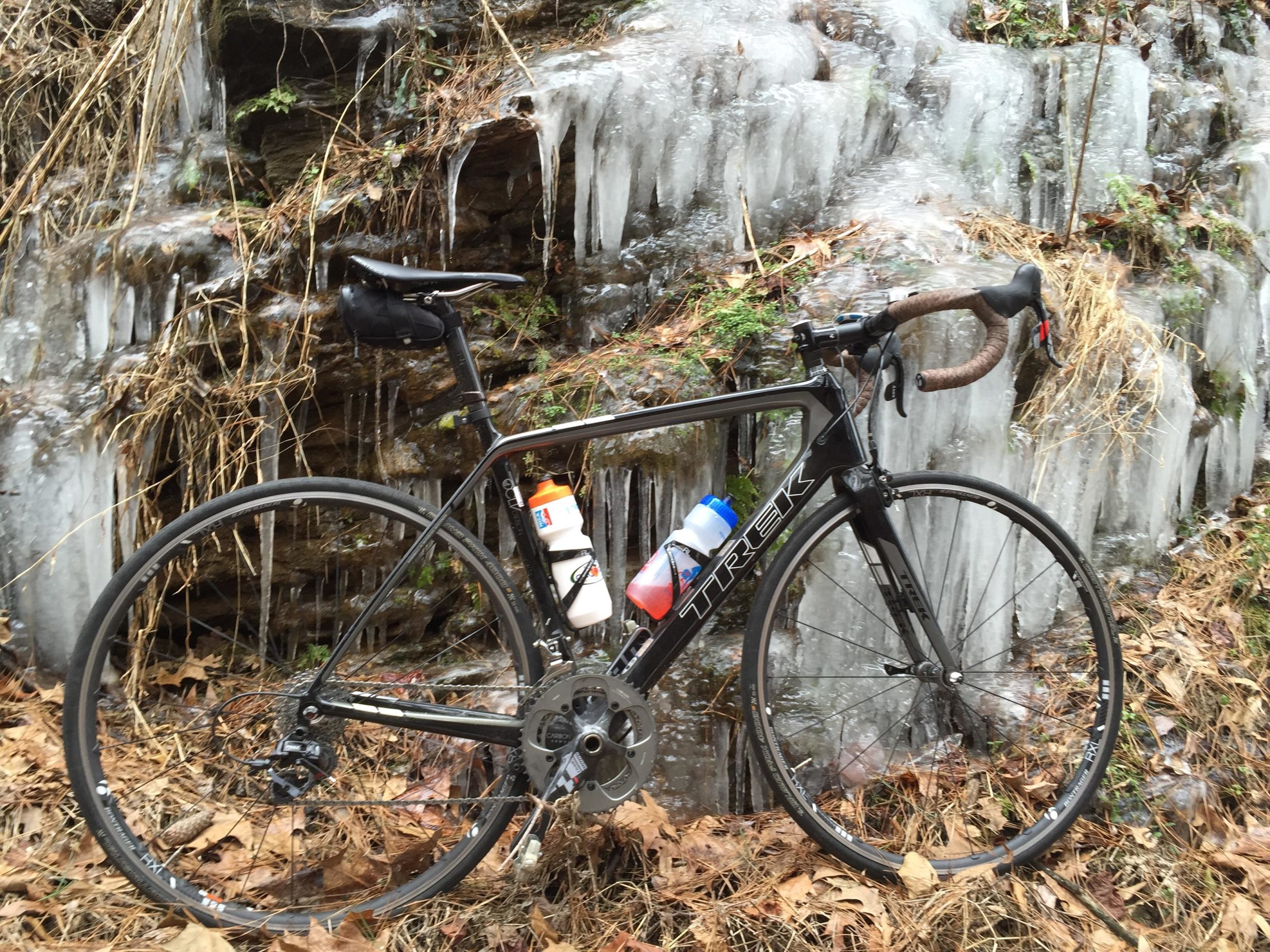 A black road bicycle with water bottles attached, positioned next to a rocky surface featuring icicles and winter foliage. The ground is covered with brown leaves, indicating a cold, wintry environment. Silver Comet Rail Trail mountain bike trail.