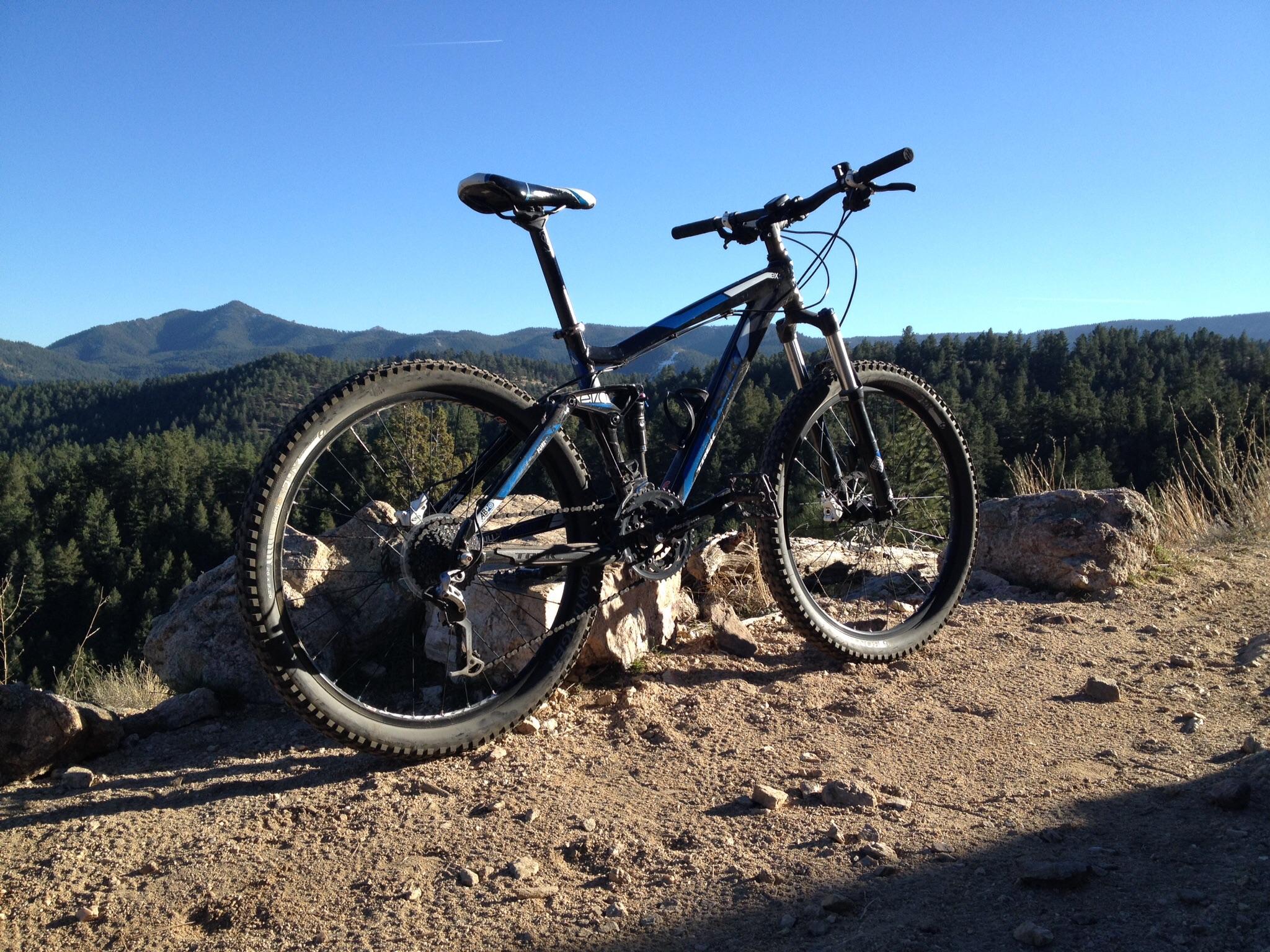 Trek Fuel EX 5: A mountain bike parked on a rocky ledge, overlooking a scenic view of rolling hills and dense forests under a clear blue sky.