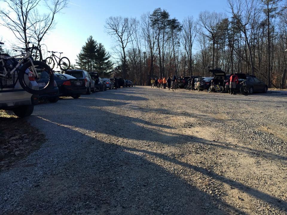 A gravel parking area filled with parked cars, some with bicycles on their rooftops. In the background, a group of people is getting ready to ride, surrounded by trees and bathed in late afternoon sunlight. Meadowood mountain bike trail.