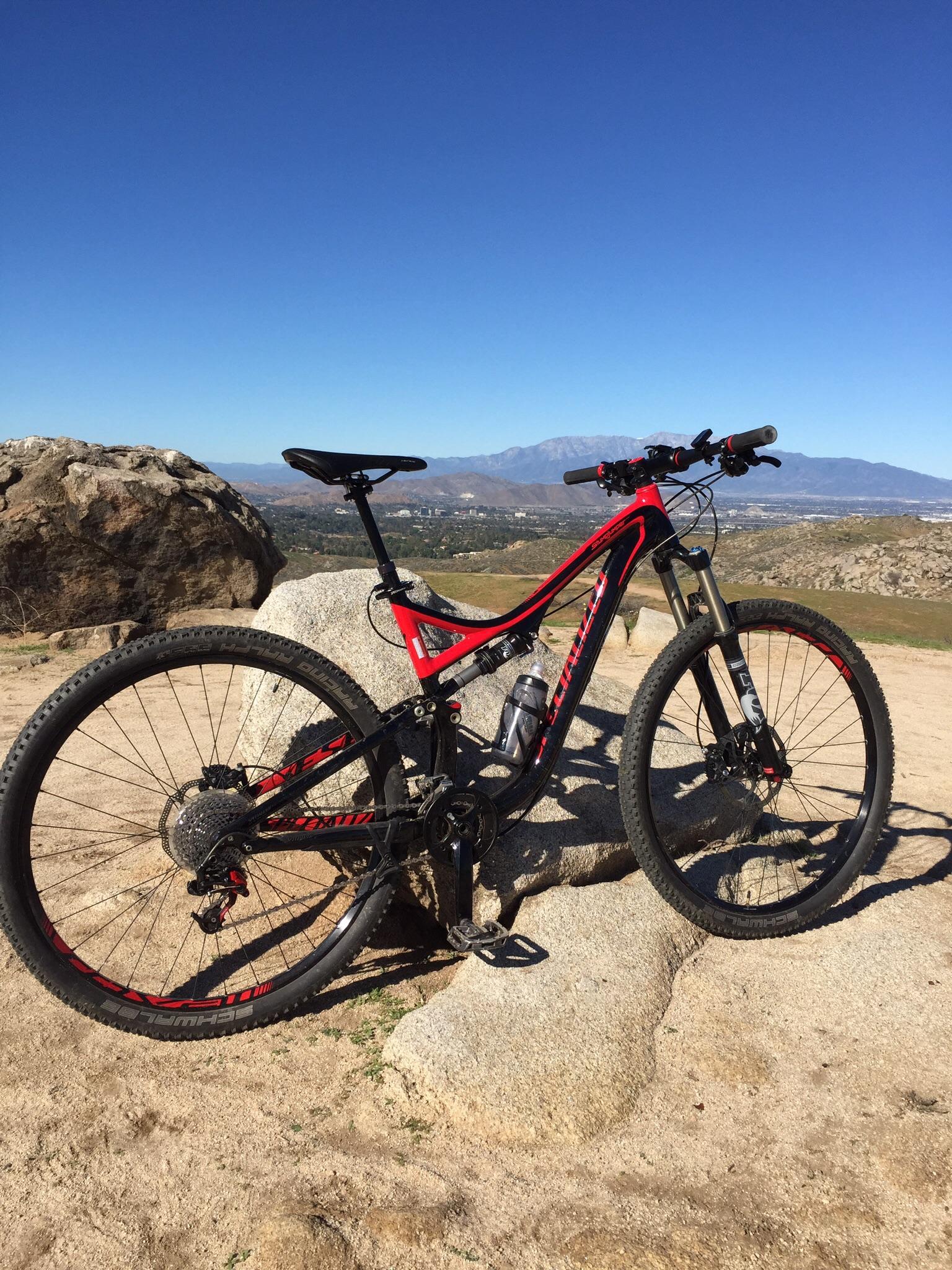 Specialized Stumpjumper FSR Comp 29er: A red and black mountain bike leaning against a large rock on a sunny day, with a clear blue sky and distant mountains in the background. The terrain is sandy and rocky, indicating an outdoor biking trail.