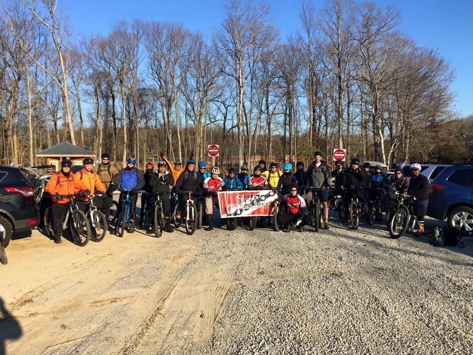 A group of mountain bikers gathered at a gravel parking area, smiling and posing for a photo. They are dressed in various biking gear, holding bikes and a large banner. Leafless trees are visible in the background and clear blue skies above. Meadowood mountain bike trail.