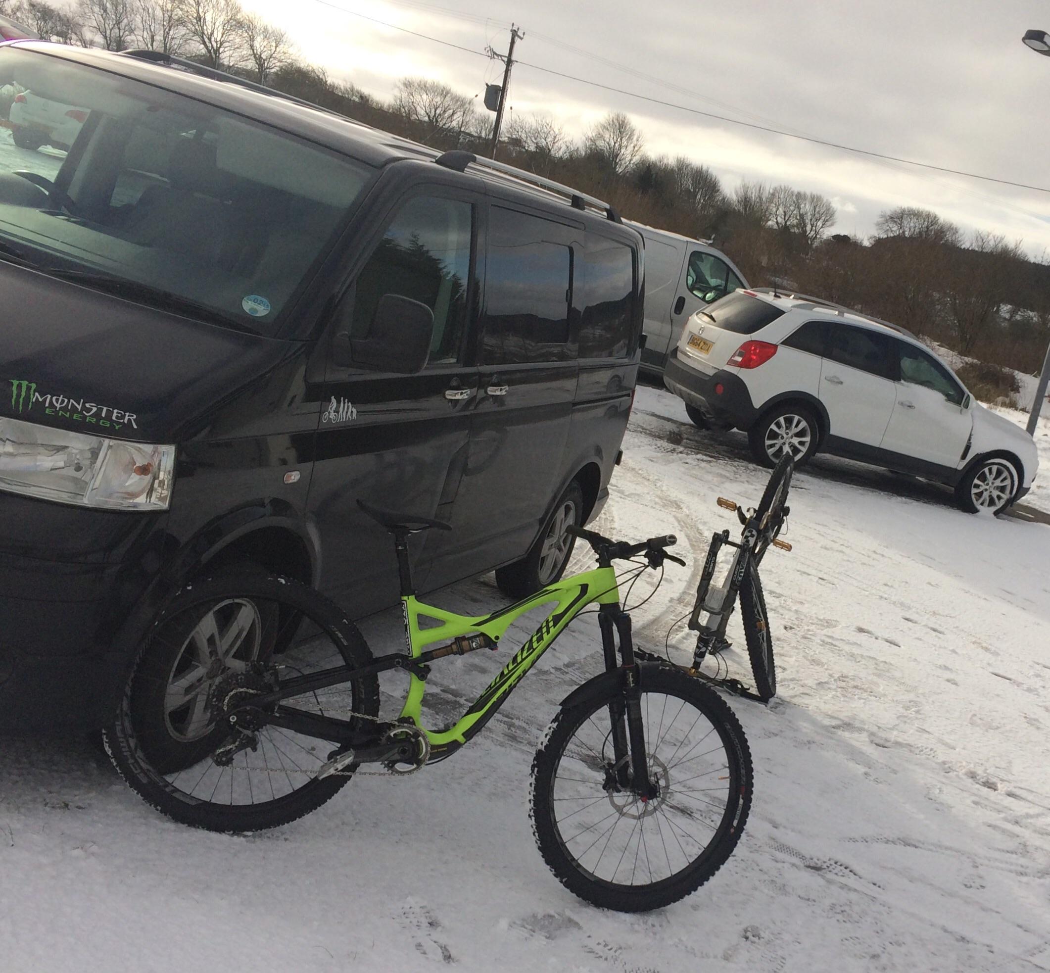 Specialized Stumpjumper FSR Expert Carbon Evo 650B: A black van parked in a snowy area, with a bright green mountain bike positioned in front of it. Another bicycle is visible in the background, and several vehicles are parked along a snowy road surrounded by trees. The sky is overcast.