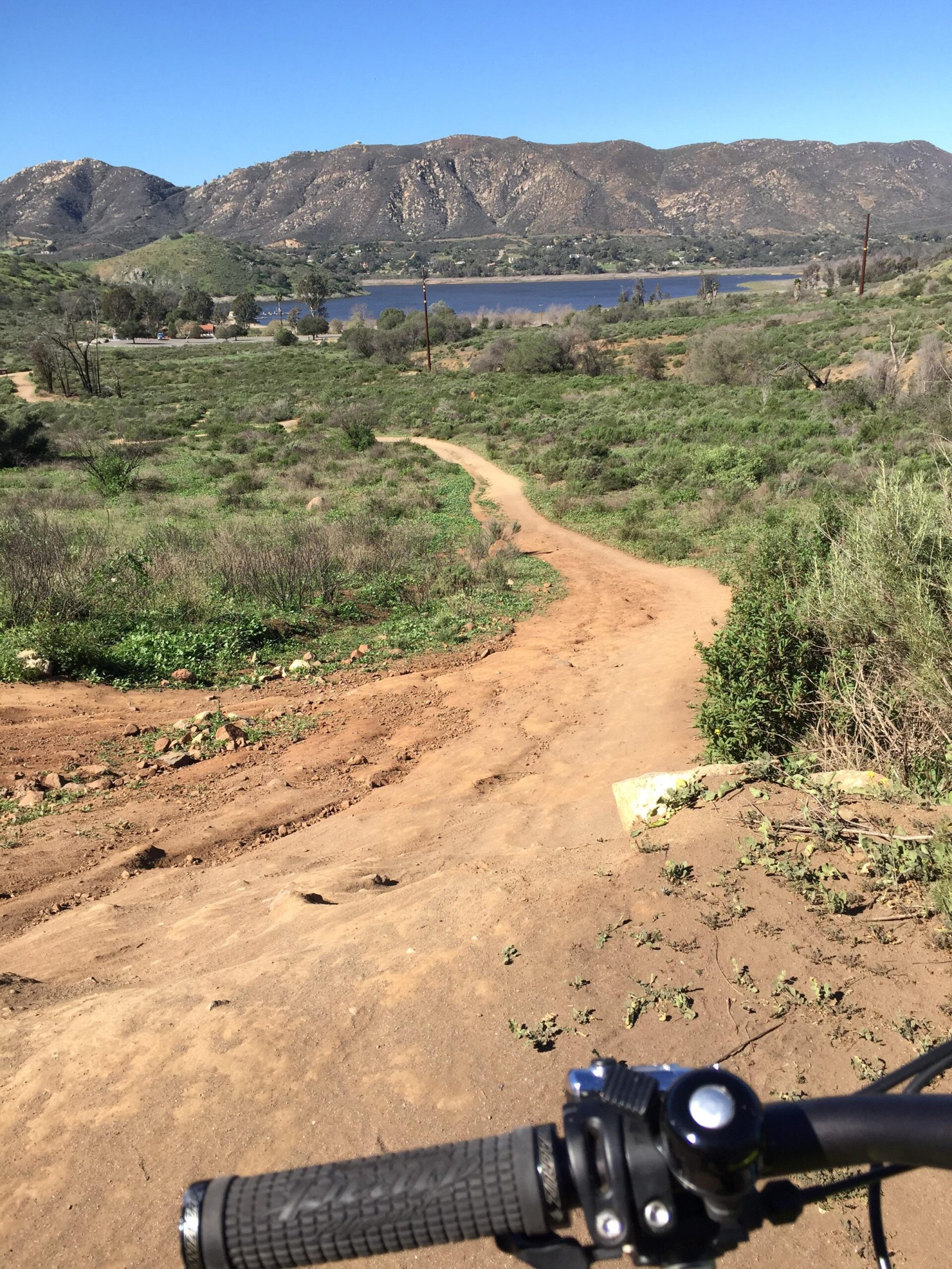 A scenic view of a dirt bike trail winding through green vegetation, leading down towards a lake surrounded by mountains under a clear blue sky. In the foreground, the handlebars of a mountain bike are visible, emphasizing the outdoor adventure setting. Lake Hodges mountain bike trail.
