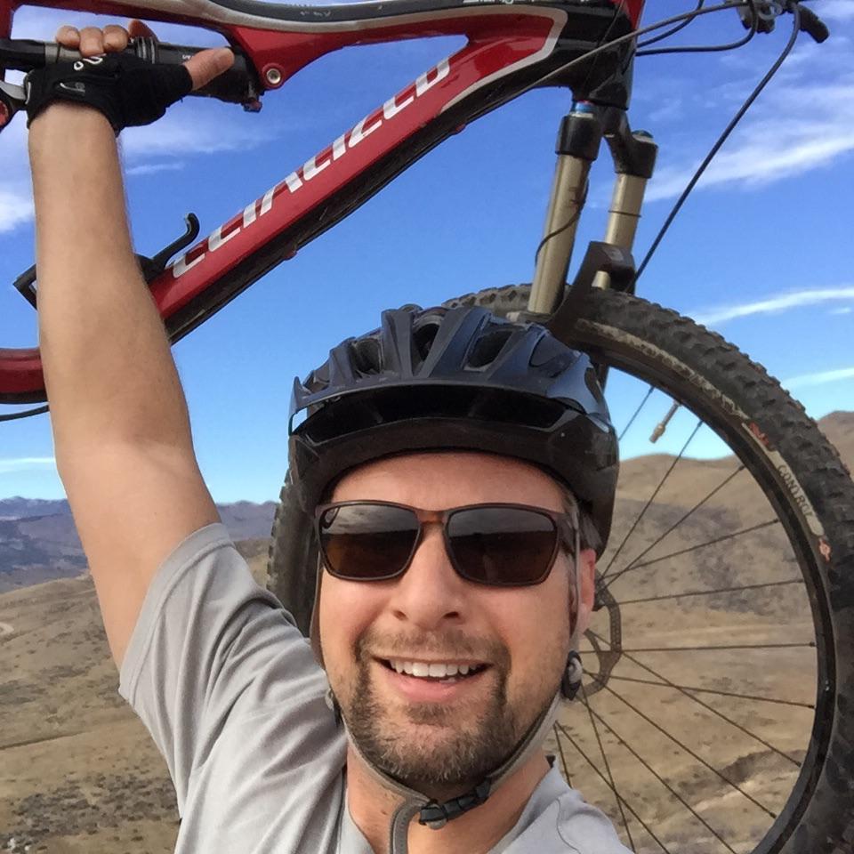 Specialized Stumpjumper FSR: A man wearing sunglasses and a helmet smiles widely while holding a mountain bike above his head in a scenic outdoor setting with hills and a blue sky in the background.