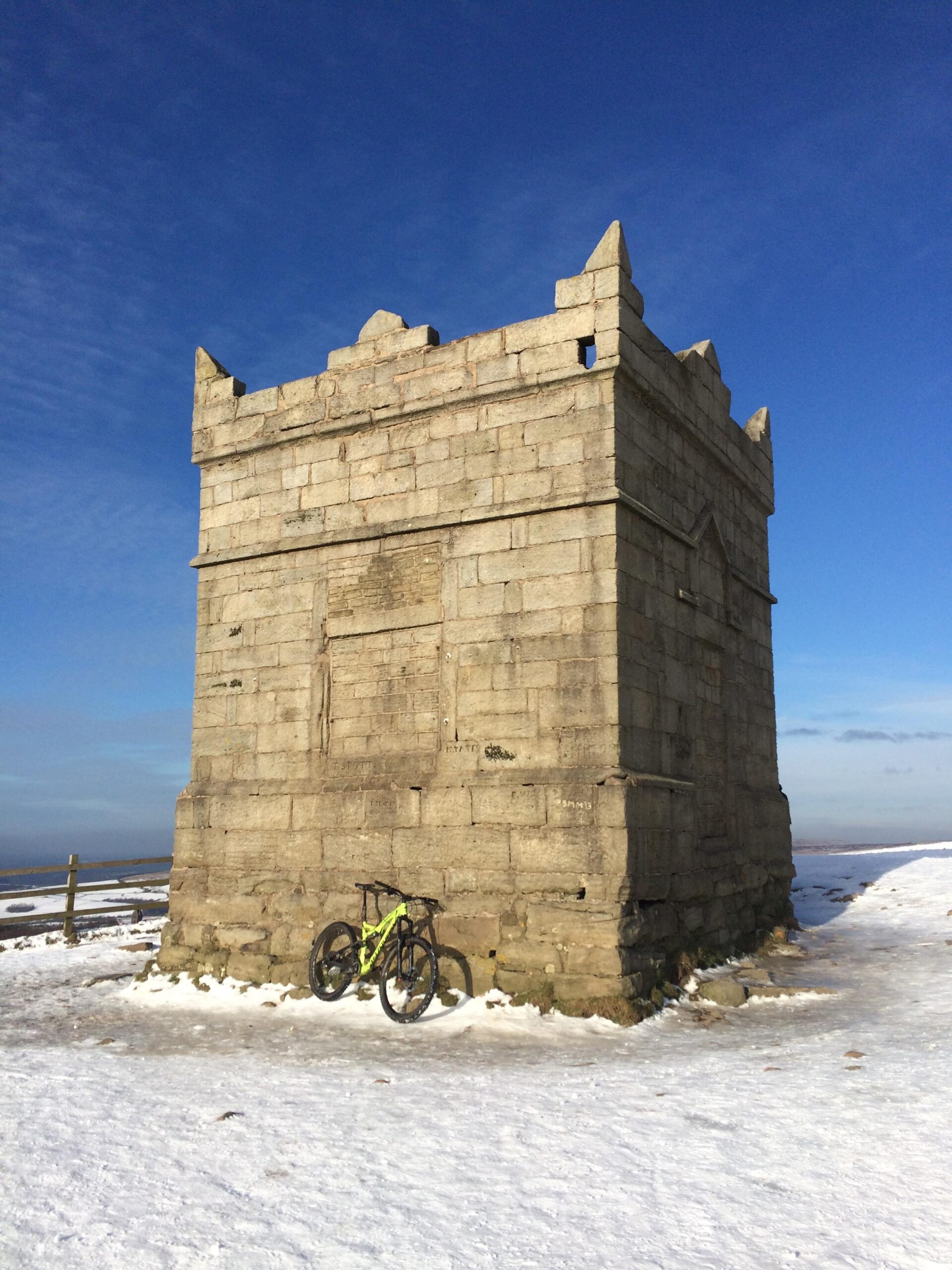 A stone tower with pointed corners stands against a clear blue sky, partially surrounded by a snowy landscape. A bright green mountain bike leans against the base of the tower, adding a pop of color to the scene. Rivington Pike mountain bike trail.