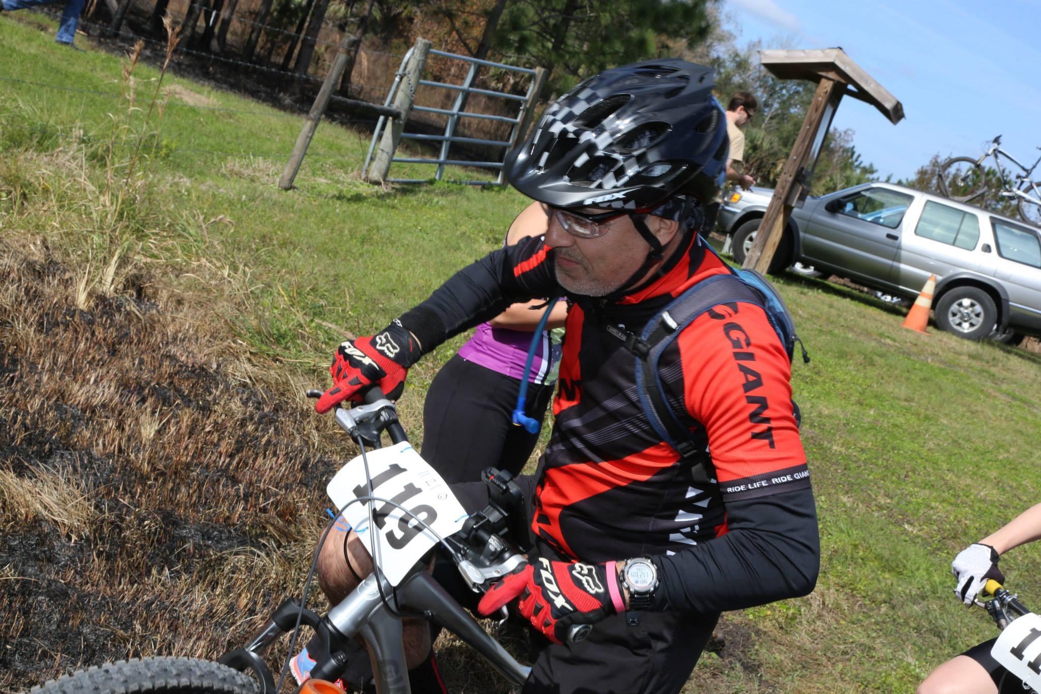 A cyclist in a red and black jersey, wearing a helmet and gloves, adjusts his bike at the starting line of a mountain biking event. In the background, a group of spectators and a parked vehicle can be seen, alongside a grassy area with some burnt vegetation. The cyclist's bib number, 119, is visible on his bike. Little Big Econ State Forest mountain bike trail.