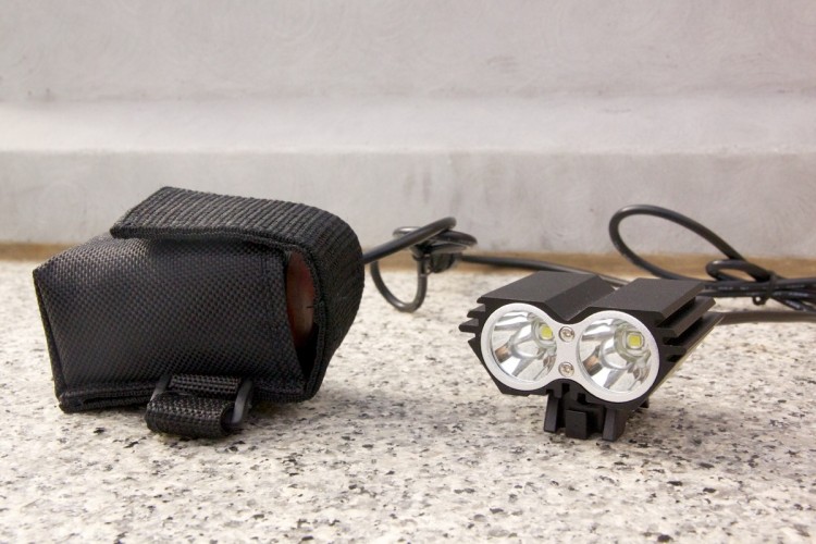 A black fabric pouch next to a dual-lens LED light mounted on a small metal heat sink, with connecting wires visible on a granite surface.