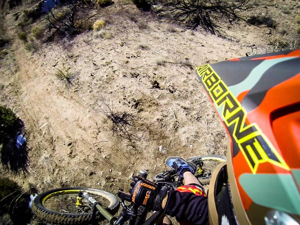 Aerial view of a mountain biker riding on a dirt trail with rugged terrain, featuring a close-up of the bike's front wheel and the rider's helmet. The surrounding landscape includes sandy soil and sparse vegetation.