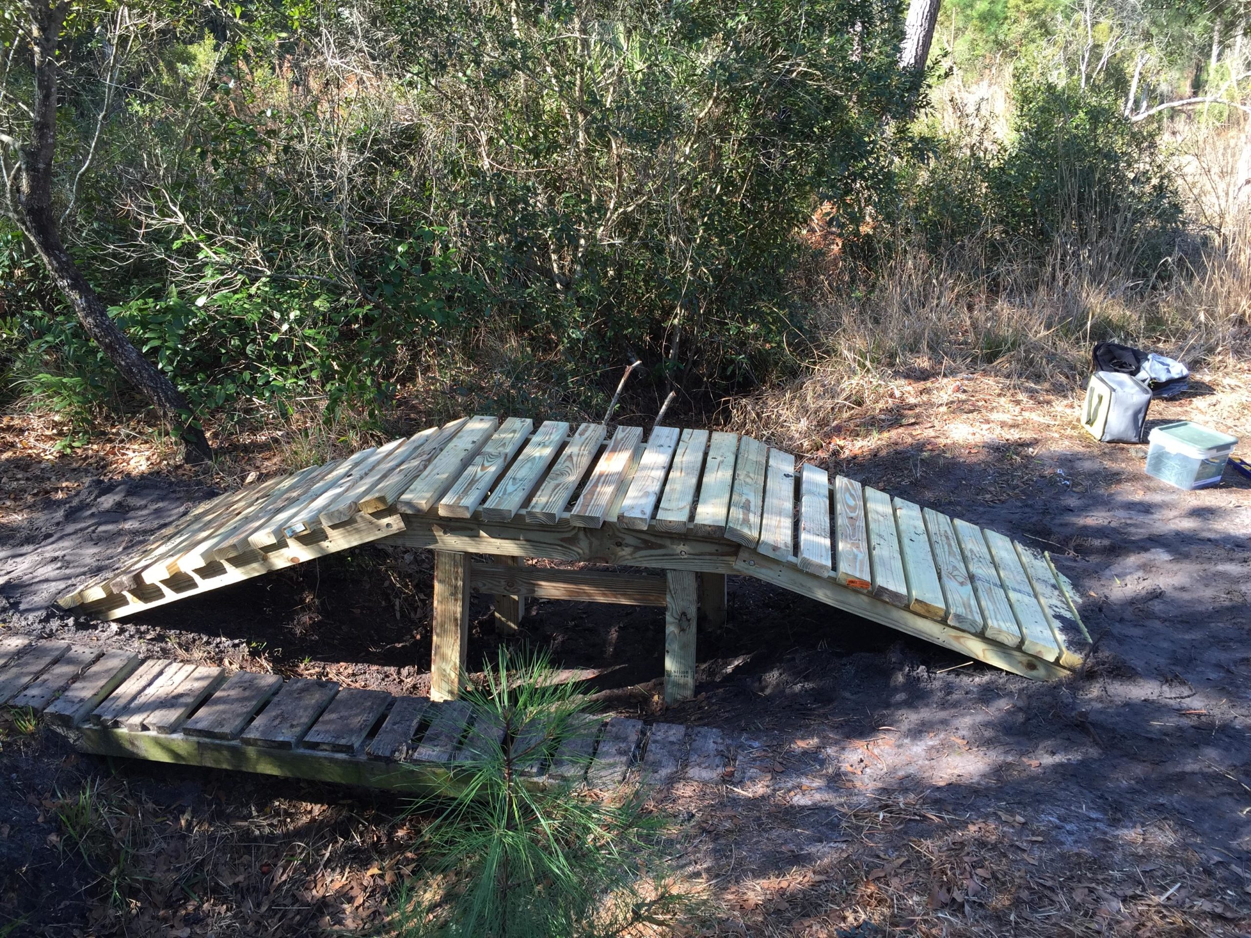 A wooden bridge structure with a sloped ramp leading into a trench, surrounded by dense brush and greenery. The scene includes some tools and equipment in the background, suggesting an area under construction or maintenance. Nocatee mountain bike trail.