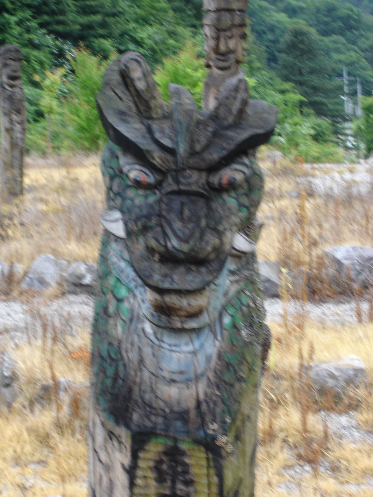 A close-up of a carved wooden statue resembling a dragon or mythical creature, featuring intricate details and vibrant colors. The statue has prominent eyes, a textured surface, and a fierce expression. In the background, other carved figures and a natural landscape of trees and dry grass are visible, suggesting an outdoor setting. The image is slightly out of focus. Machesan mountain bike trail.