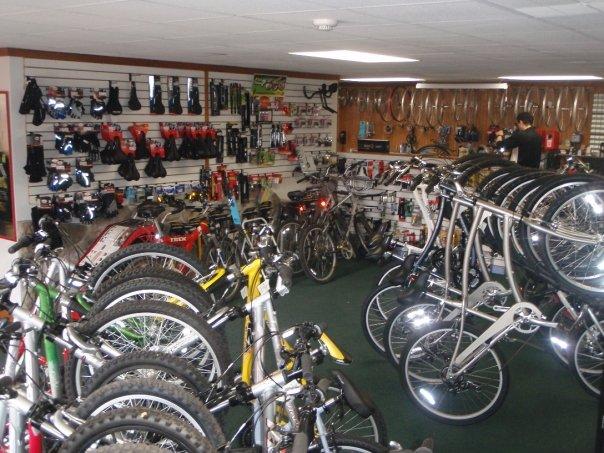 A bike shop interior featuring a variety of bicycles, including mountain bikes and road bikes, displayed on shelves and racks. The store is stocked with cycling accessories such as helmets and gloves along the walls, while a customer is browsing in the background.