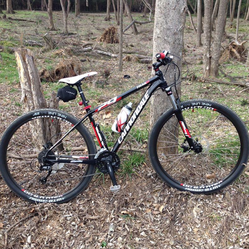 A black and red mountain bike leaning against a tree in a forested area, with a water bottle attached to the frame and a small bag mounted on the seat post. The background features a mix of grass and fallen branches typical of a wooded environment. Augusta Canal mountain bike trail.
