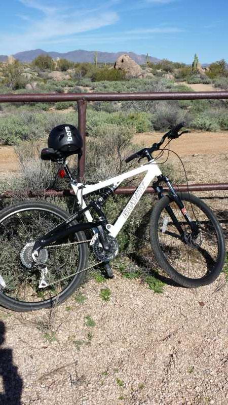 A black and white mountain bike is leaning against a rustic metal fence in a desert landscape. In the background, there are rocky formations and sparse vegetation under a clear blue sky. A black helmet is mounted on the bike's handlebars. Pima Road and Dynamite Blvd mountain bike trail.