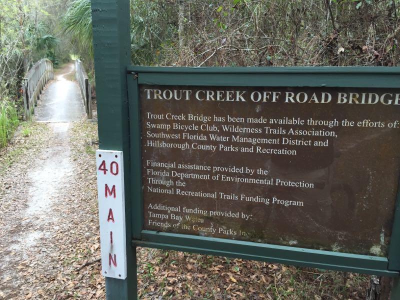 Alt text: A sign at the Trout Creek Off Road Bridge, detailing the contributions of various organizations and funding sources for the bridge's construction. The path is visible leading to the bridge, surrounded by dense vegetation. Flatwoods Park mountain bike trail.