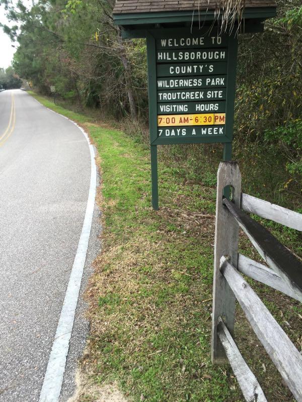 Welcome sign for Hillsborough County's Wilderness Park, Trout Creek site, displaying visiting hours from 7:00 AM to 6:30 PM, seven days a week, with a road and wooden fence in the foreground. Wilderness Trails Park Loop mountain bike trail.