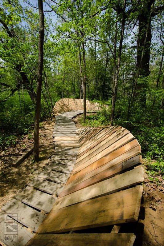 A winding wooden pathway through a lush green forest, surrounded by trees and sunlight filtering through the leaves. The path features an elevated, curving design, showcasing a combination of wooden planks and supports. Outback Trail at Imagination Glenn mountain bike trail.