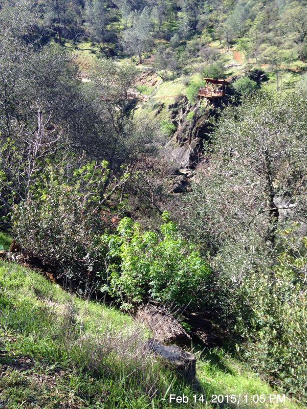 A scenic view of a lush green landscape, featuring dense vegetation, trees, and rocky cliffs. In the distance, a wooden structure can be seen perched on the edge of the cliff, surrounded by nature. The image captures the tranquility and beauty of a natural environment. Hidden Falls Regional Park mountain bike trail.