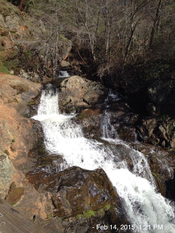 A cascading waterfall flowing over rocks, surrounded by trees in a natural setting. The image captures the movement of the water as it tumbles down, with sunlight reflecting off the surface. Hidden Falls Regional Park mountain bike trail.
