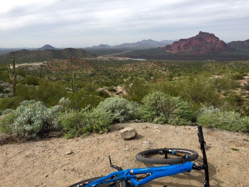 A panoramic view of a southwestern landscape featuring rolling hills and red rock formations under a cloudy sky. In the foreground, a blue mountain bike rests on a gravel path surrounded by desert vegetation, including cacti and shrubs. The distant mountains create a scenic backdrop for the natural terrain. Hawes Loop mountain bike trail.