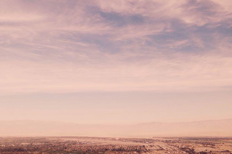 A panoramic view of a hazy landscape under a soft, cloud-filled sky, showcasing a distant mountain range. In the foreground, patches of greenery and urban development are visible, hinting at a blend of nature and city life. The Goat Trails mountain bike trail.
