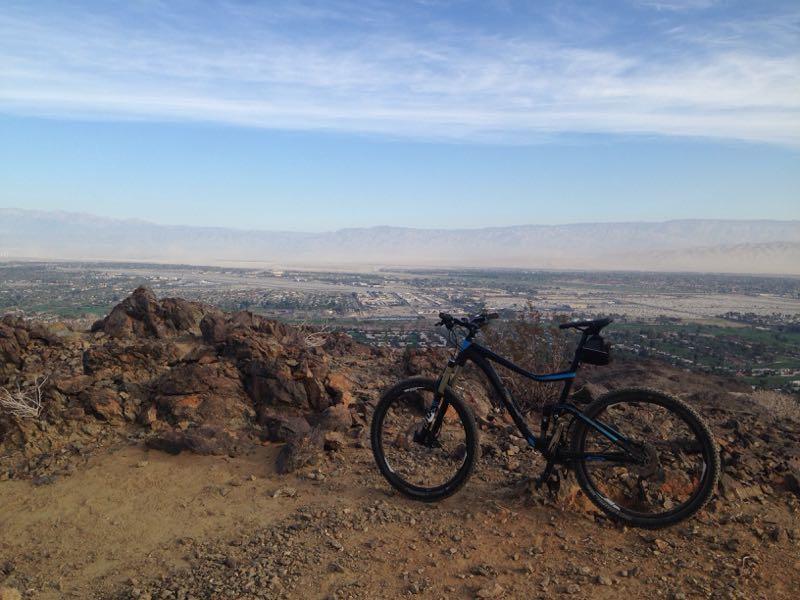 A mountain bike is resting on rocky terrain with a panoramic view of a valley below, showcasing a blend of urban and natural landscapes under a clear sky. The Goat Trails mountain bike trail.