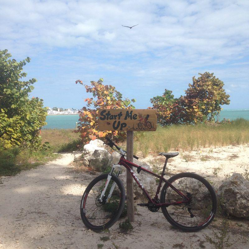 A red mountain bike leaning against a wooden sign that reads "Start Me Up...LOL," surrounded by sandy terrain and lush greenery. In the background, a tranquil body of water and a cloudy blue sky are visible. Virginia Key North Point mountain bike trail.